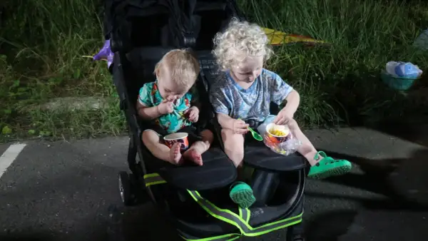 Two young children sit in a double stroller, eating corn from cups.