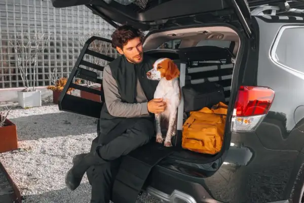 Man and dog sitting in the open trunk of a car with a dog crate and luggage.