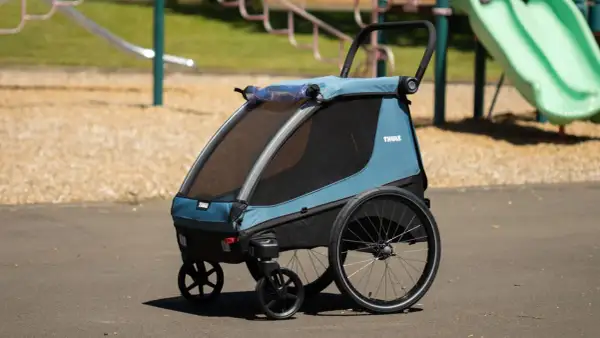 Blå Thule Courier barnvagn med stora hjul står på en asfalterad yta vid en lekplats.