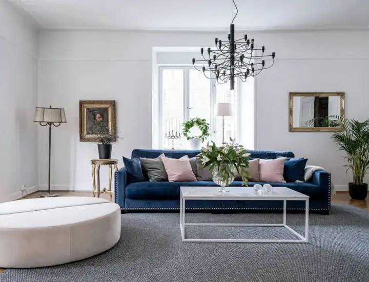 Blå soffa inspiration: En elegant salong med en djupblå sammetssoffa, prydd med kuddar i rosa och grått. Ett modernt soffbord och en stor golvlampa kompletterar rummet.